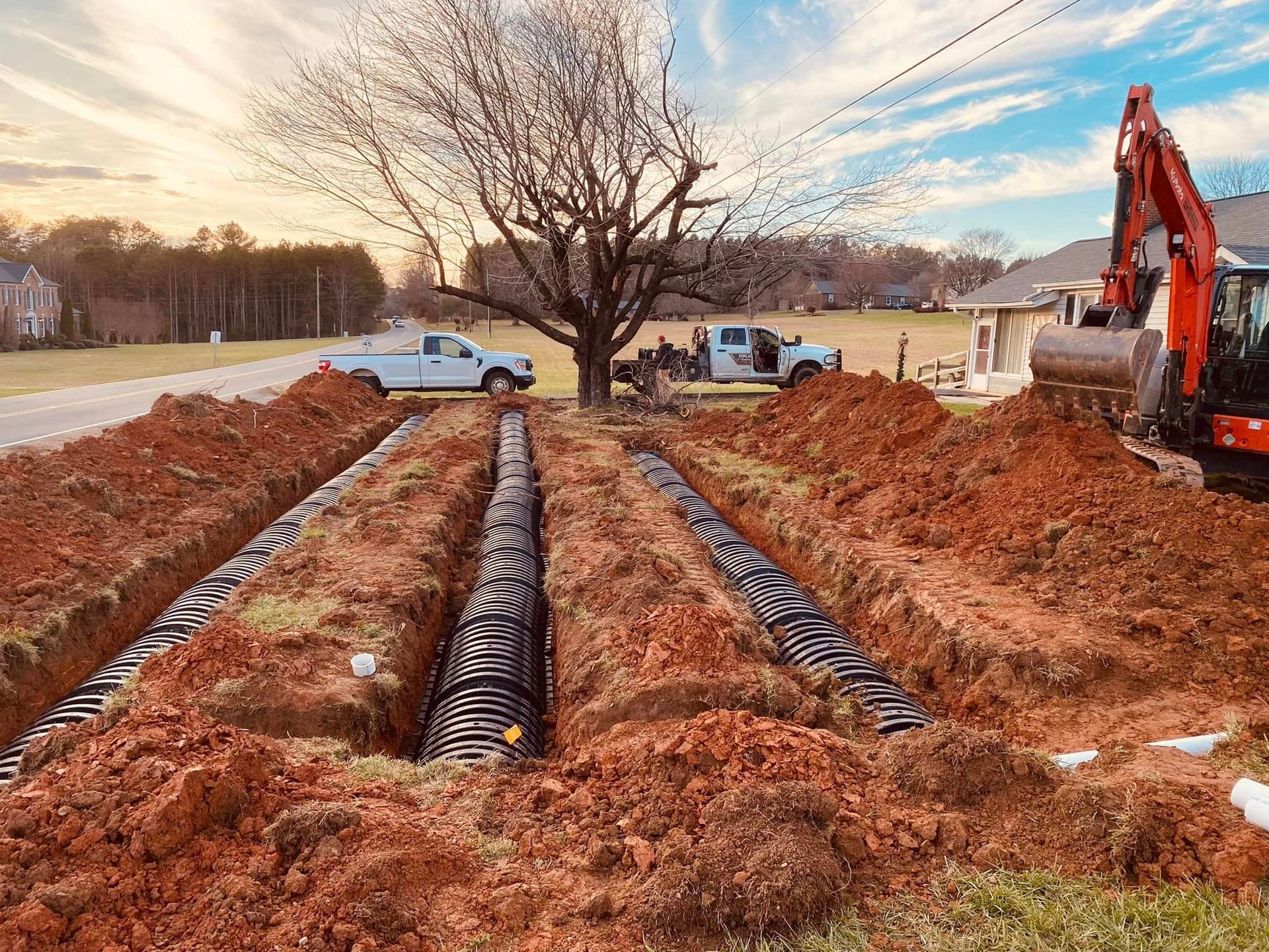 Excavation trench with black piping, dirt piles, backhoe, and workers near houses at sunset