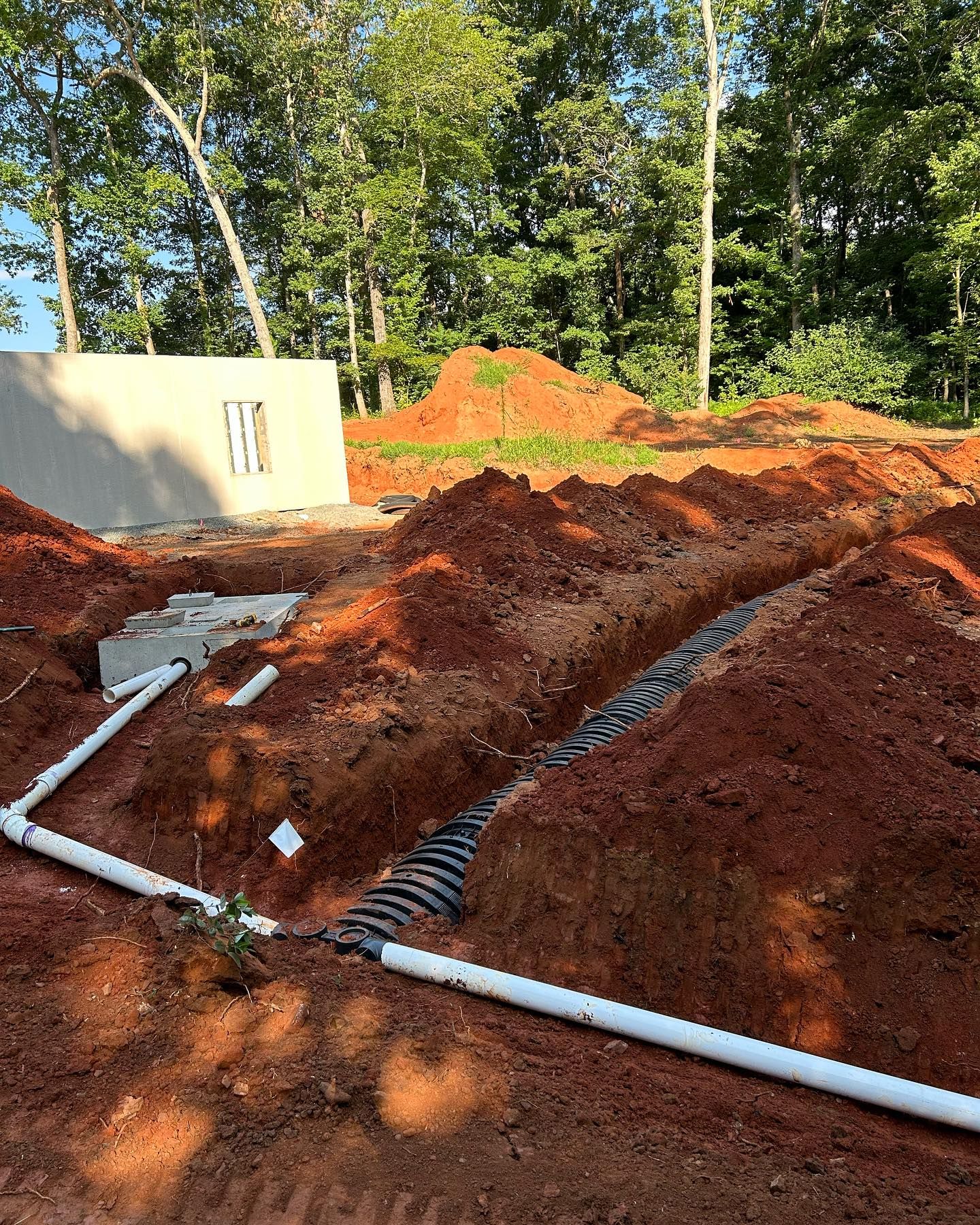Construction site with red dirt mounds, white pipes, and a concrete wall in a wooded area.