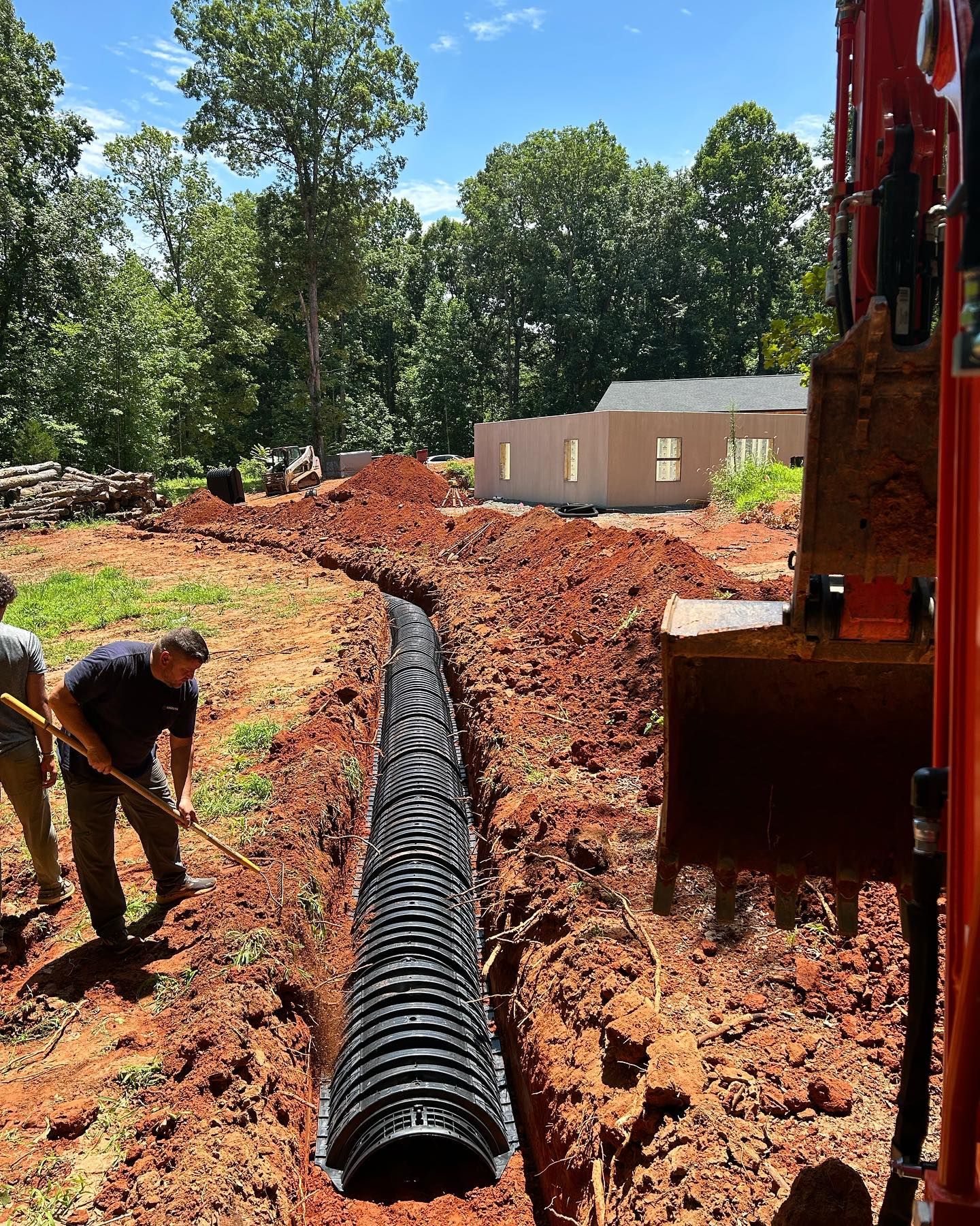 Construction trench with black drainage pipe being installed by workers beside heavy equipment on a dirt site