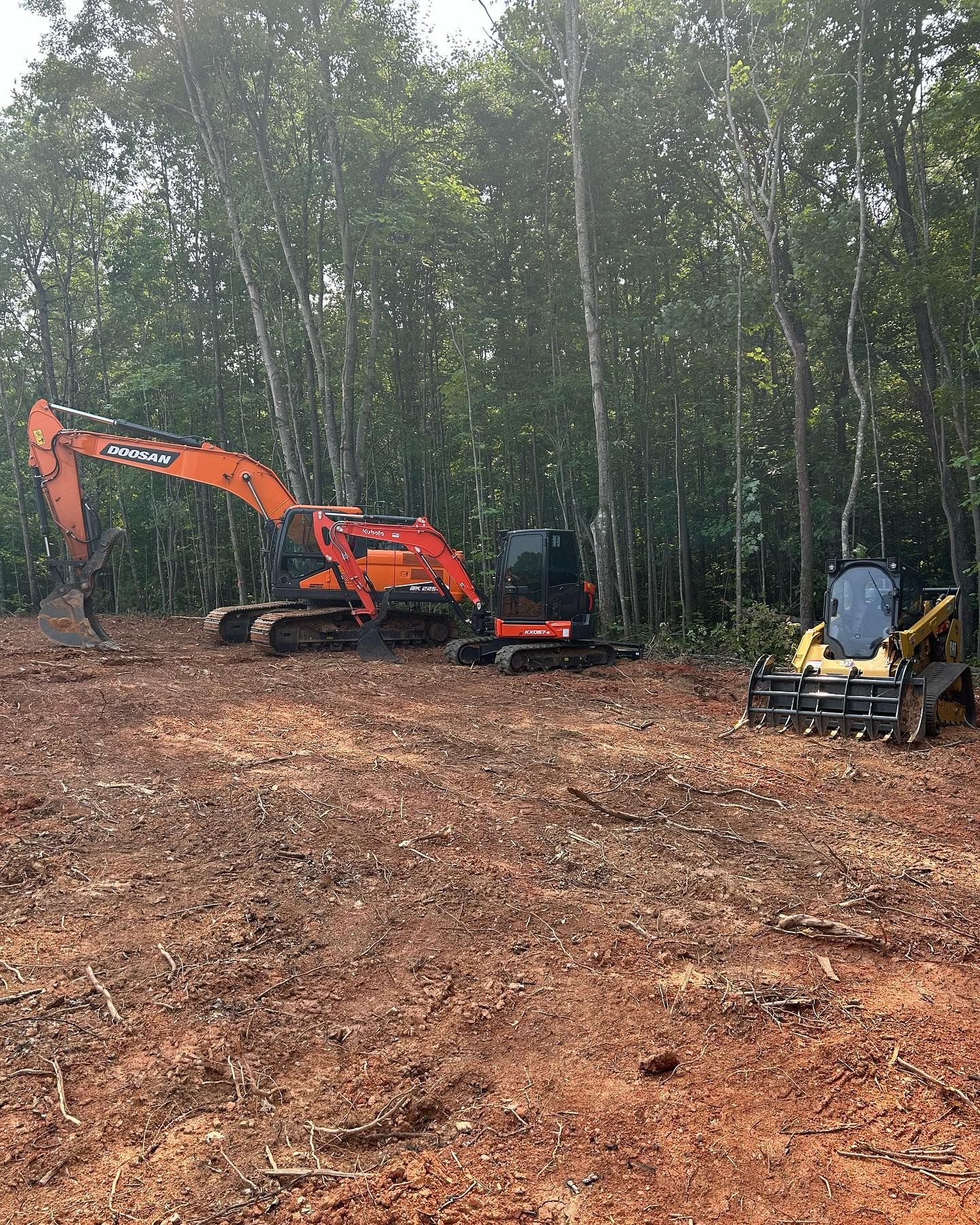 Orange excavator and skid steer on a cleared dirt lot beside a wooded tree line.