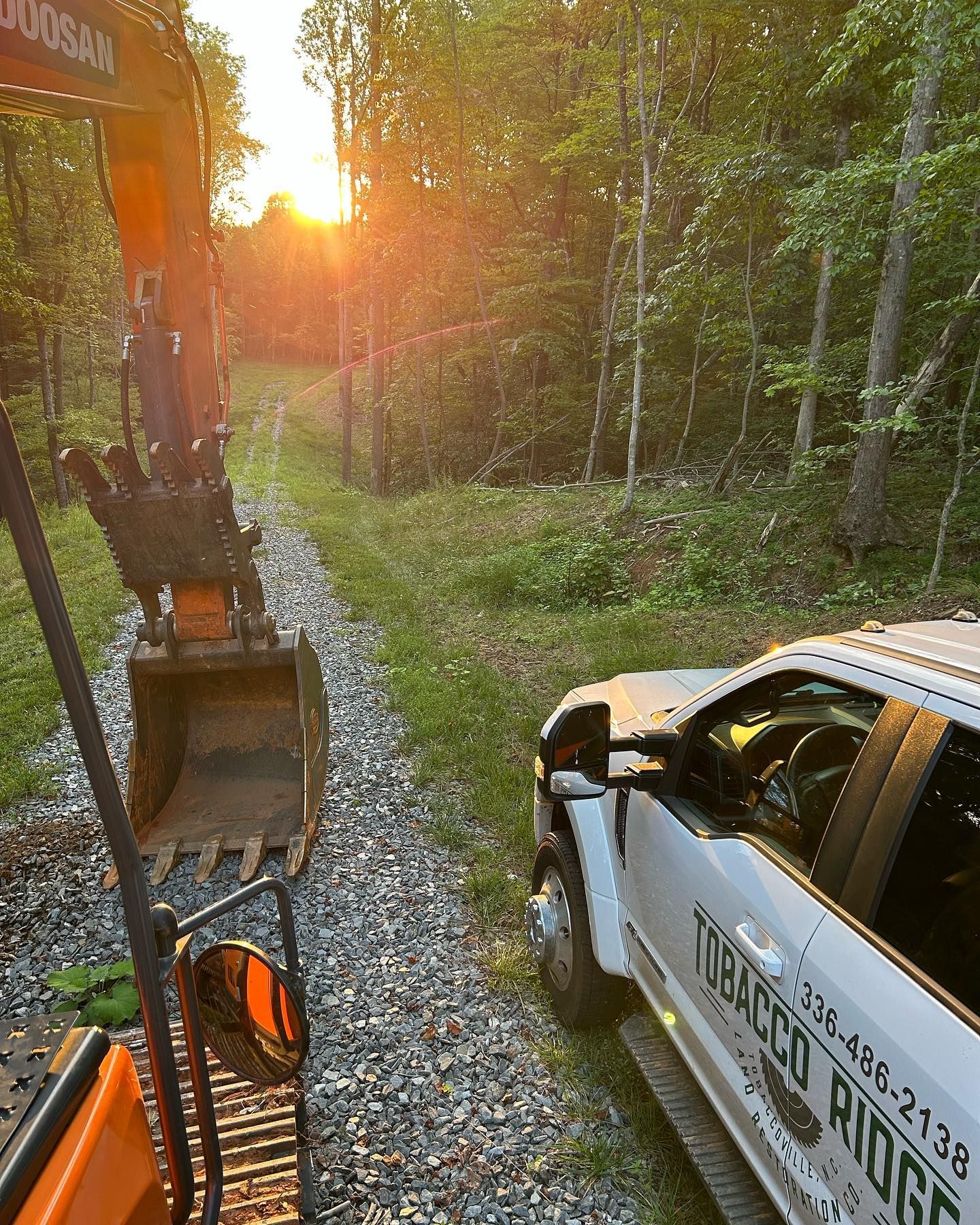 Excavator beside a white utility truck on a gravel forest road at sunset