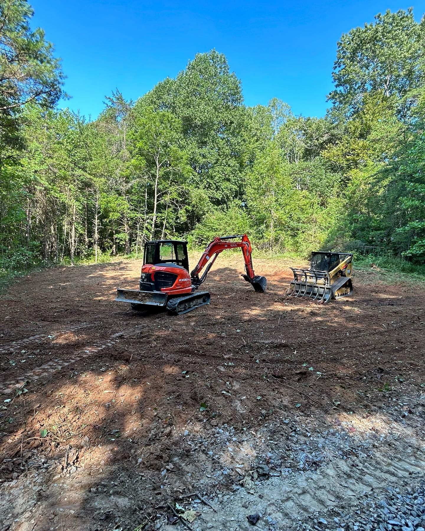 Small excavator and skid steer clearing dirt in a wooded clearing under blue sky.