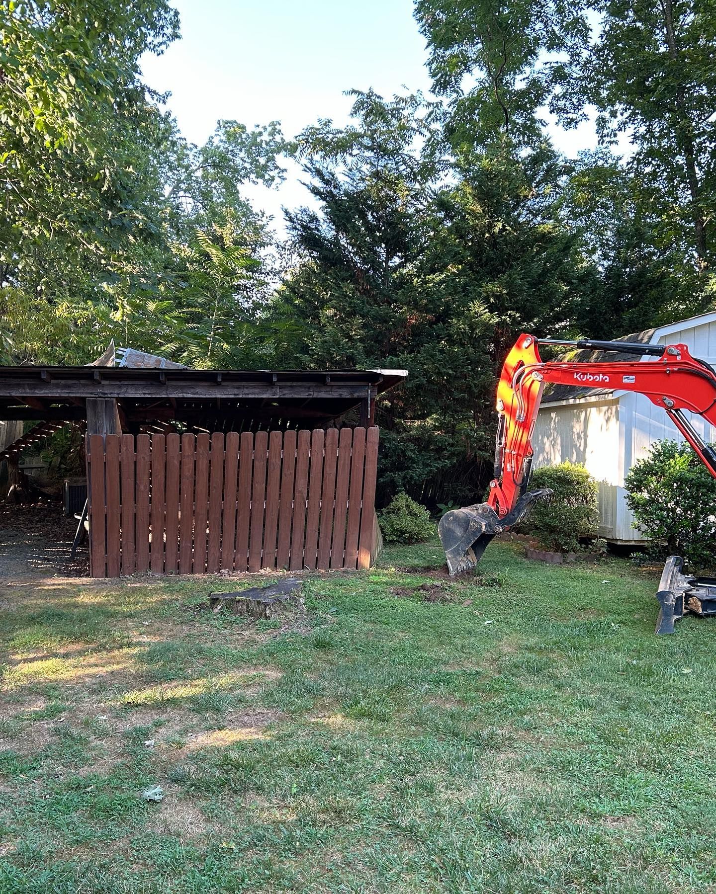 Orange excavator digging beside a wooden shed in a grassy backyard with trees