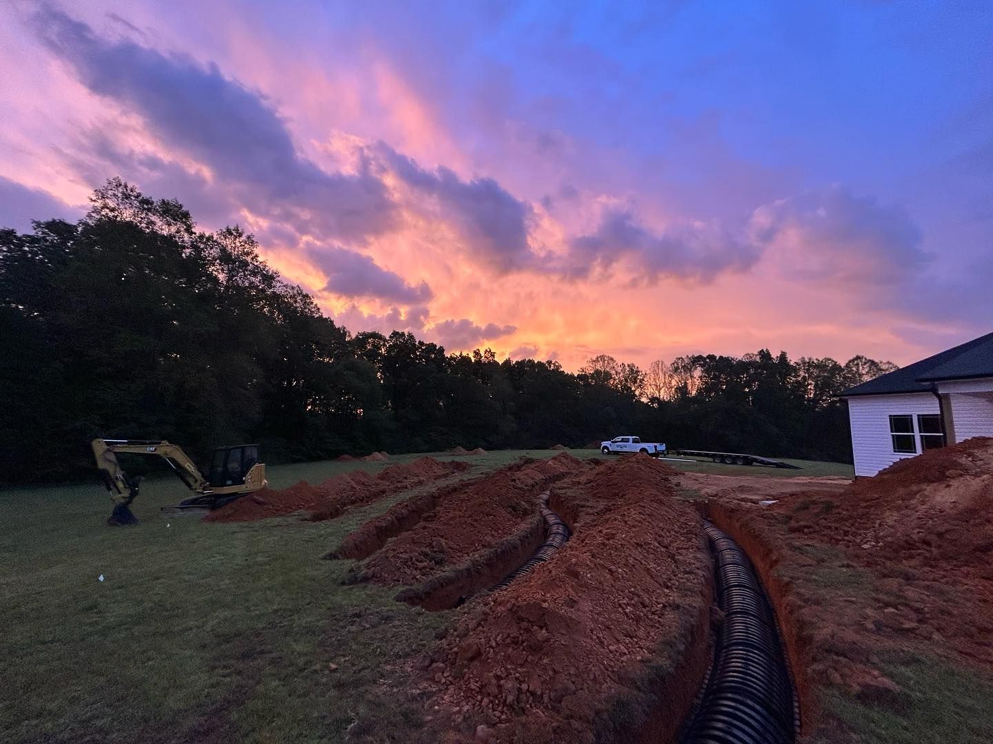 Construction trench at sunset with red dirt, an excavator, and trees silhouetted against a pink sky