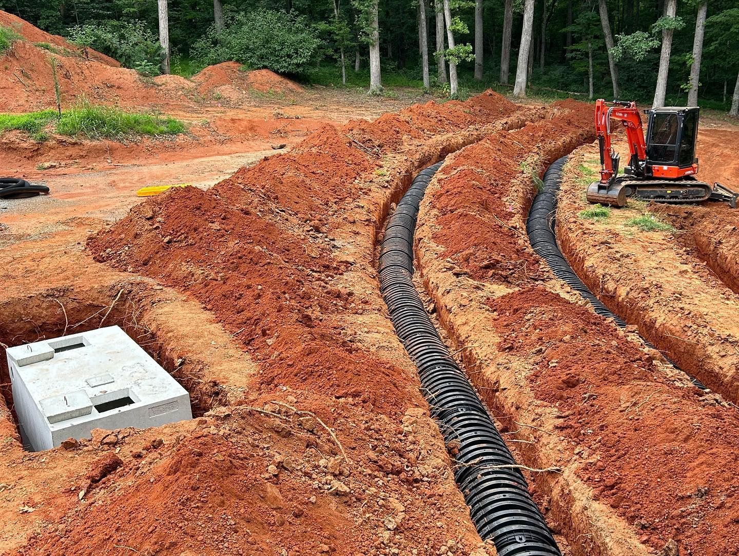 Red dirt construction site with curved black drainage pipes and a small excavator near the treeline
