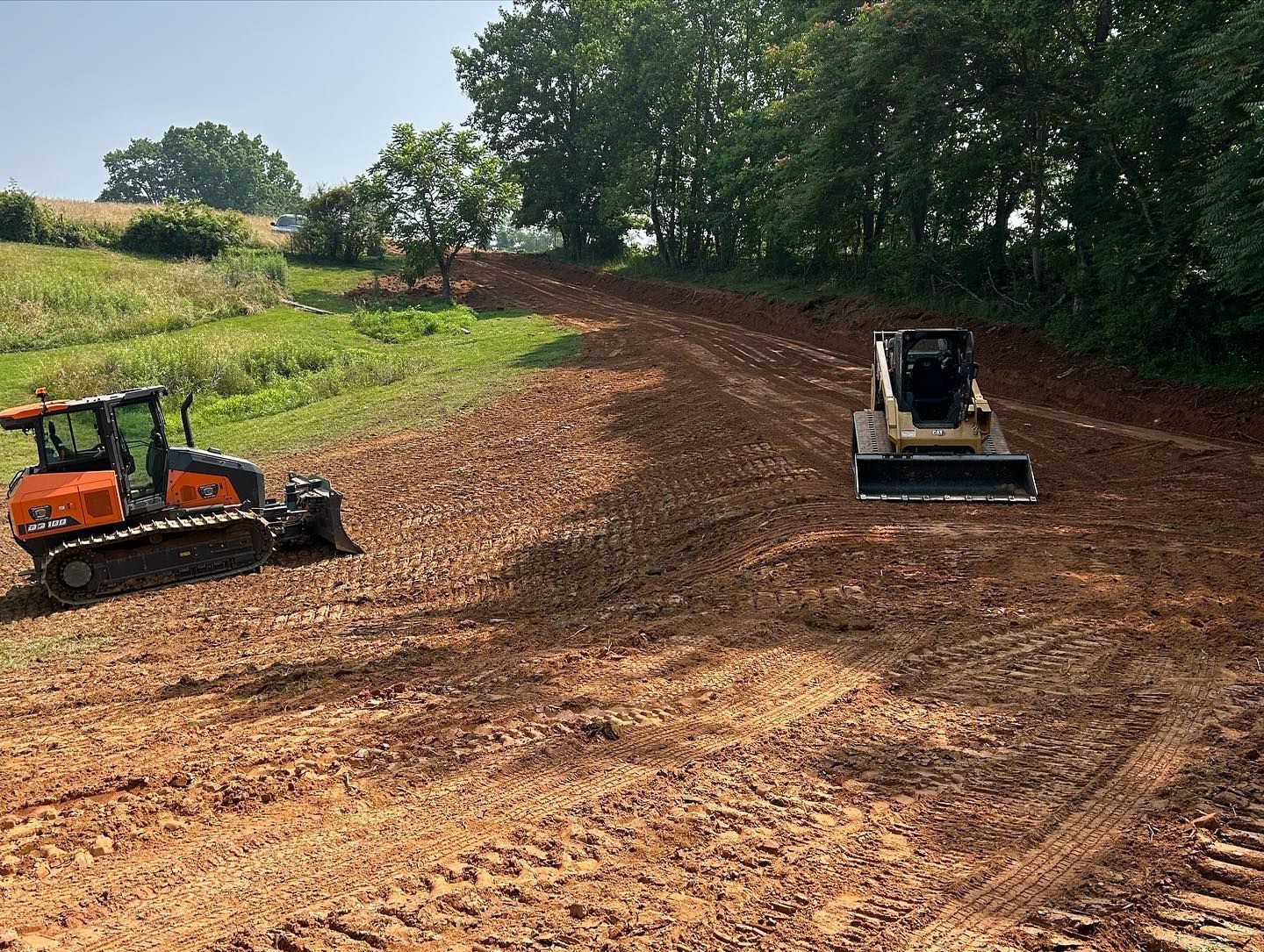 Two skid steer loaders grading a dirt lot under trees in a rural field