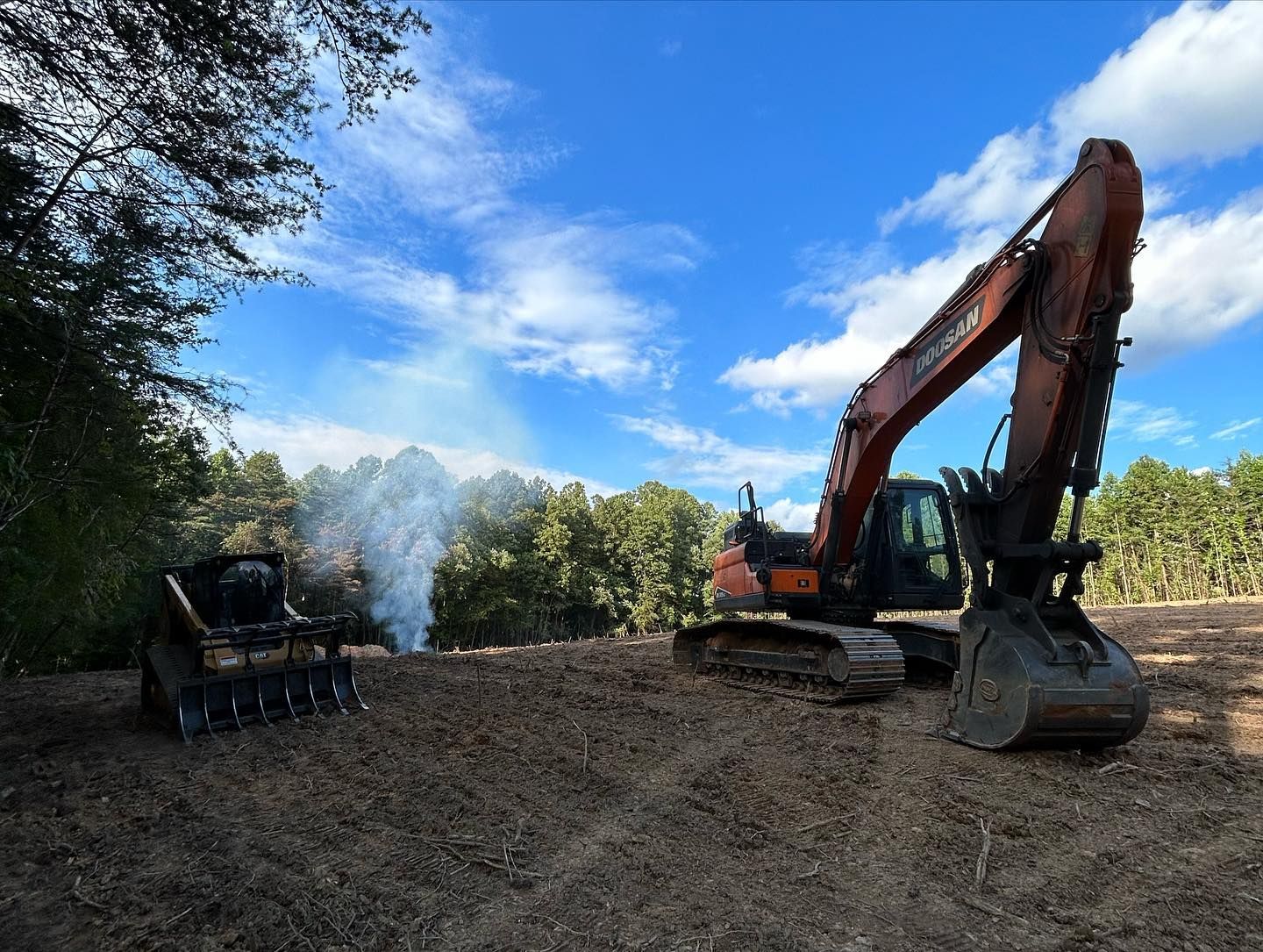 Excavator clearing a dirt lot, with smoke rising under a blue sky and trees in the background
