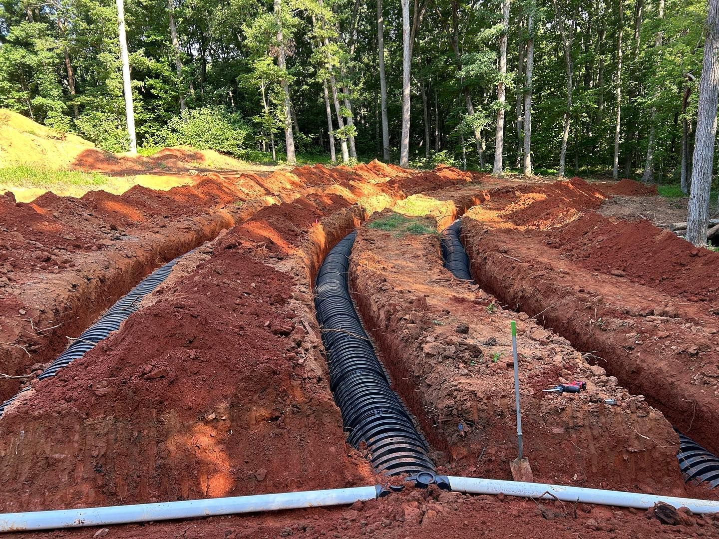 Trenches in red soil with black drainage pipes laid in a wooded construction site.