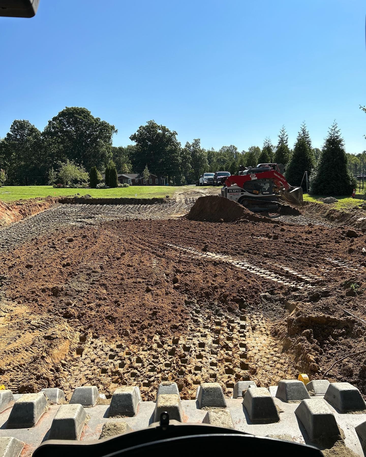 Construction site with red excavator leveling dirt beside a grassy field under a clear blue sky