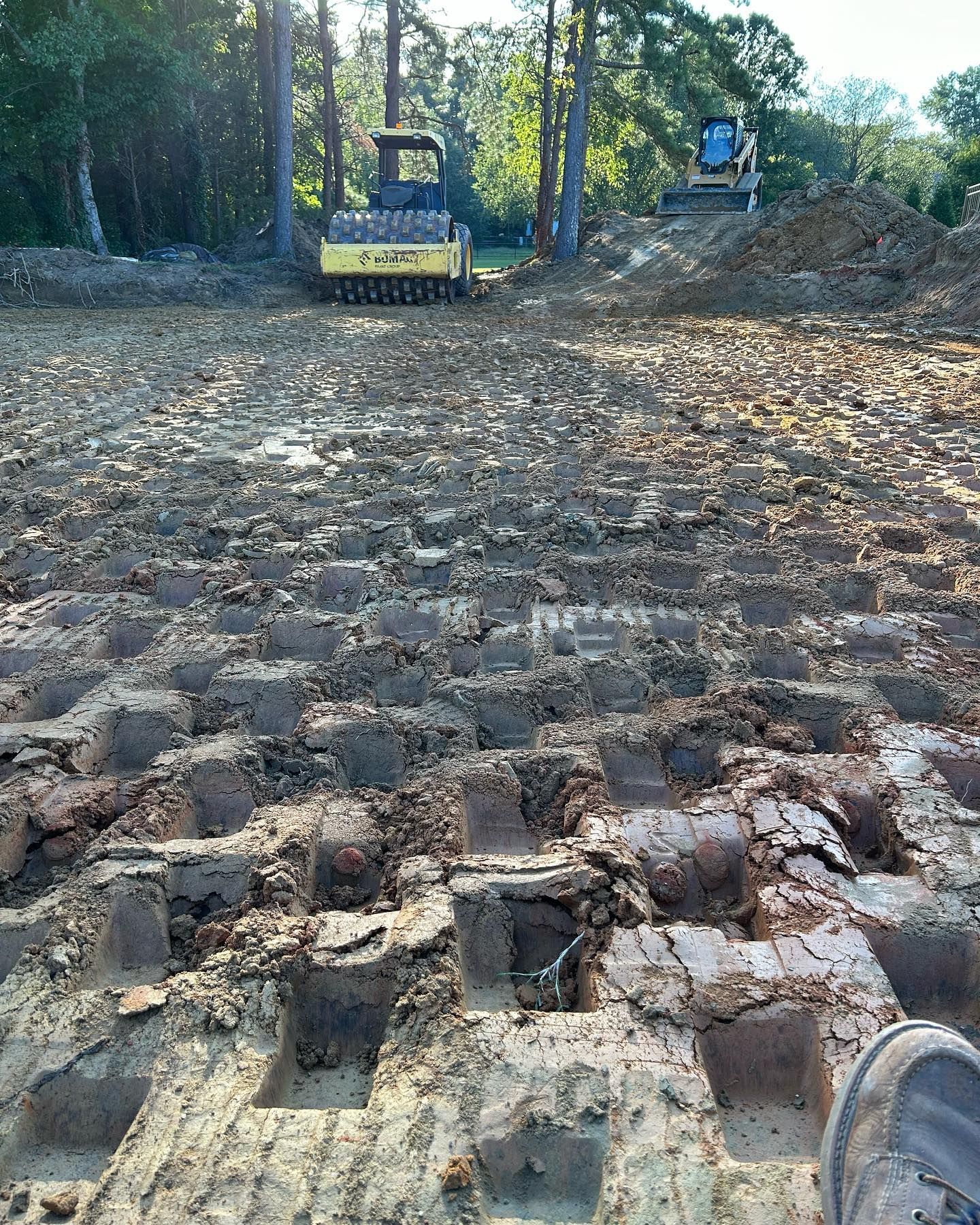 Bulldozer and excavator clearing a dirt road in a wooded area, with rough soil in the foreground
