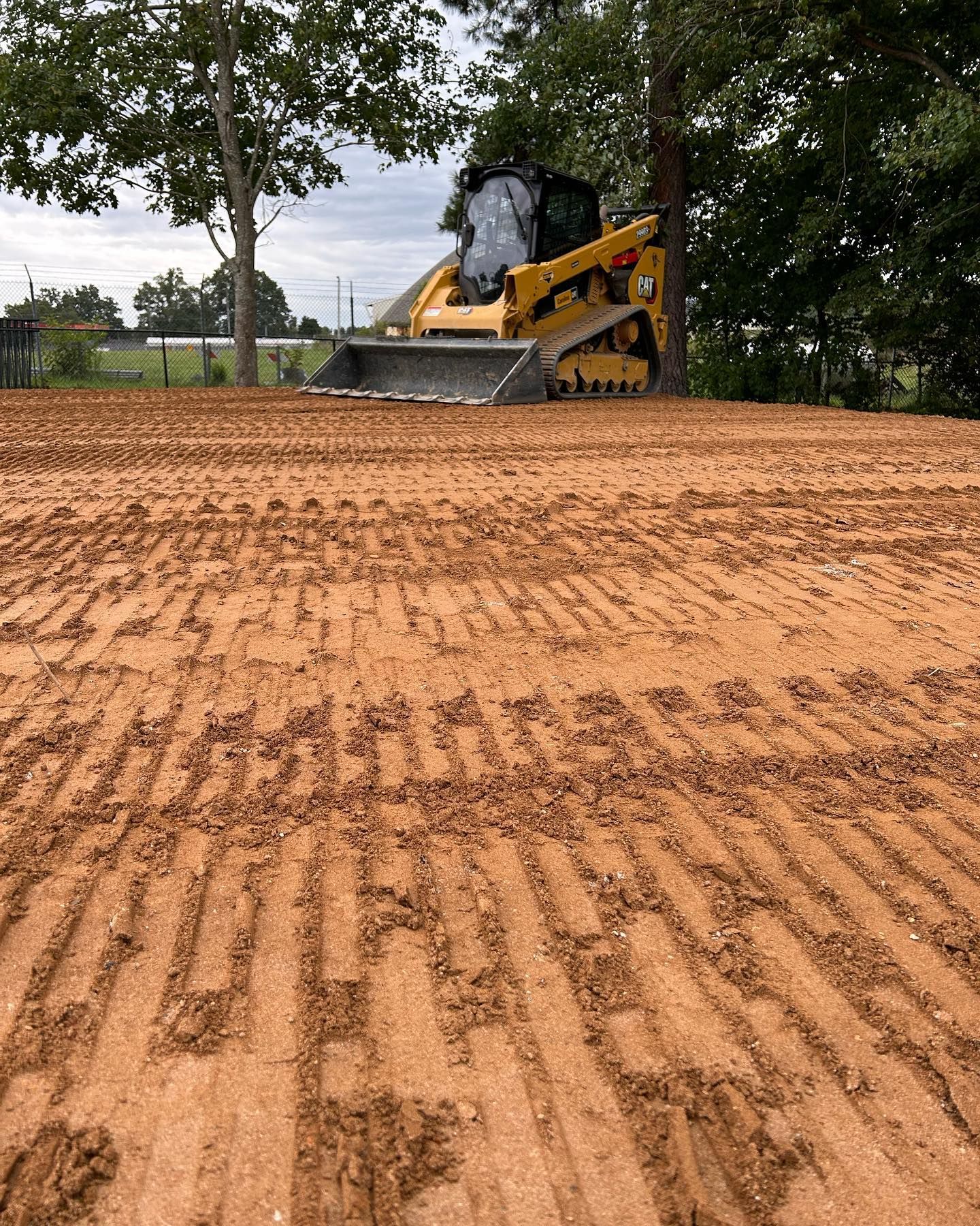 Yellow skid steer on a dirt hill near trees, grading a cleared construction site.