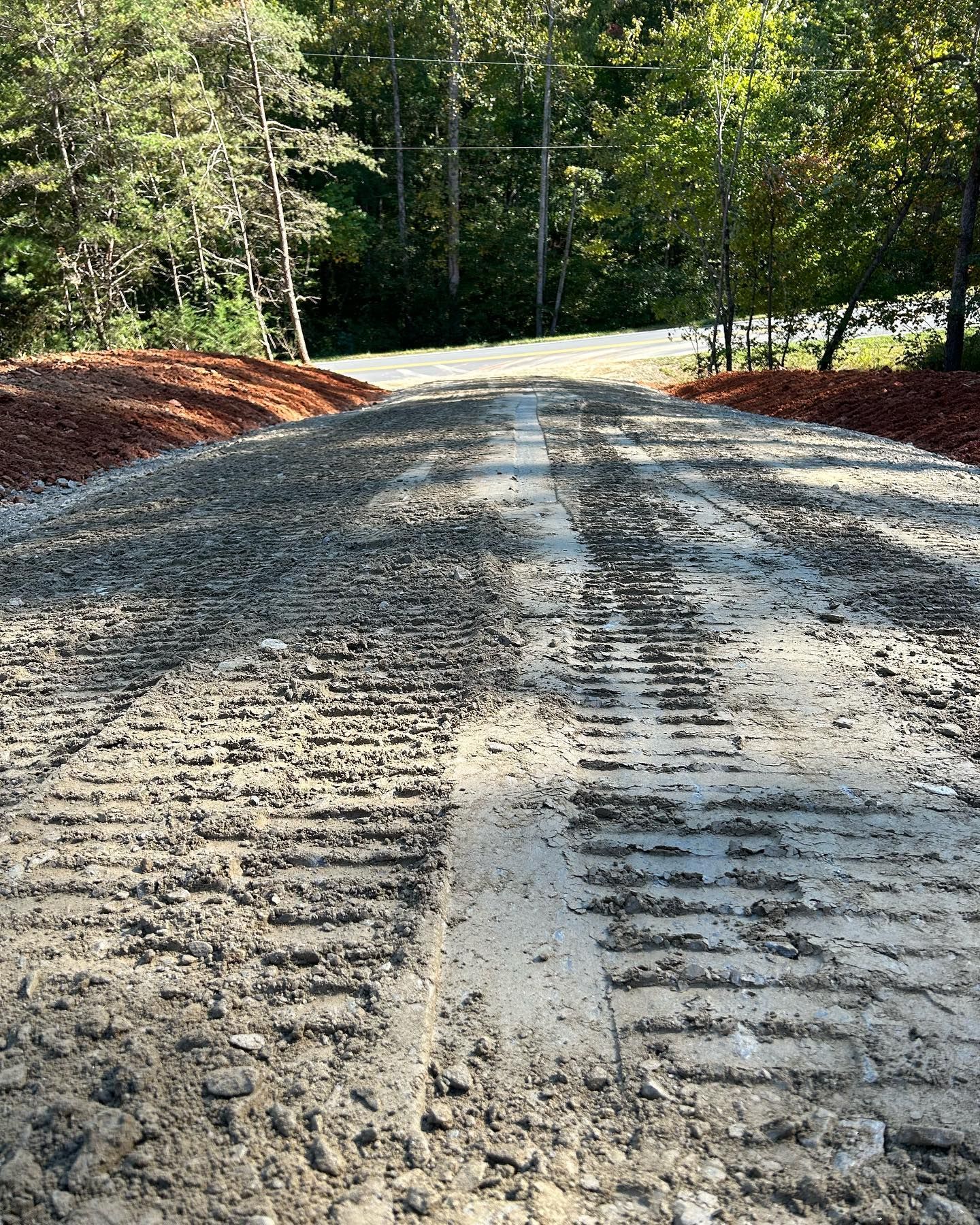 Graded gravel road through wooded area, with tire tracks and red dirt shoulders