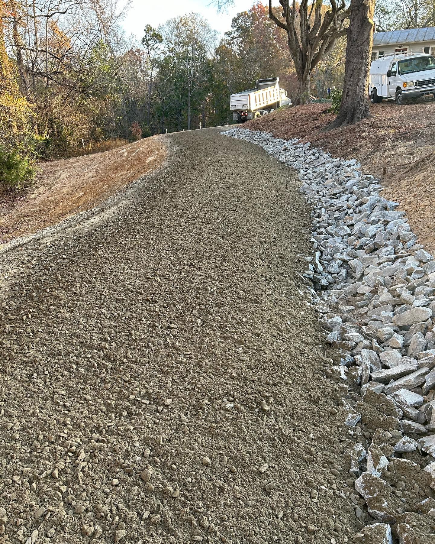 Gravel path beside a rock-lined slope in a wooded residential area with houses in the background