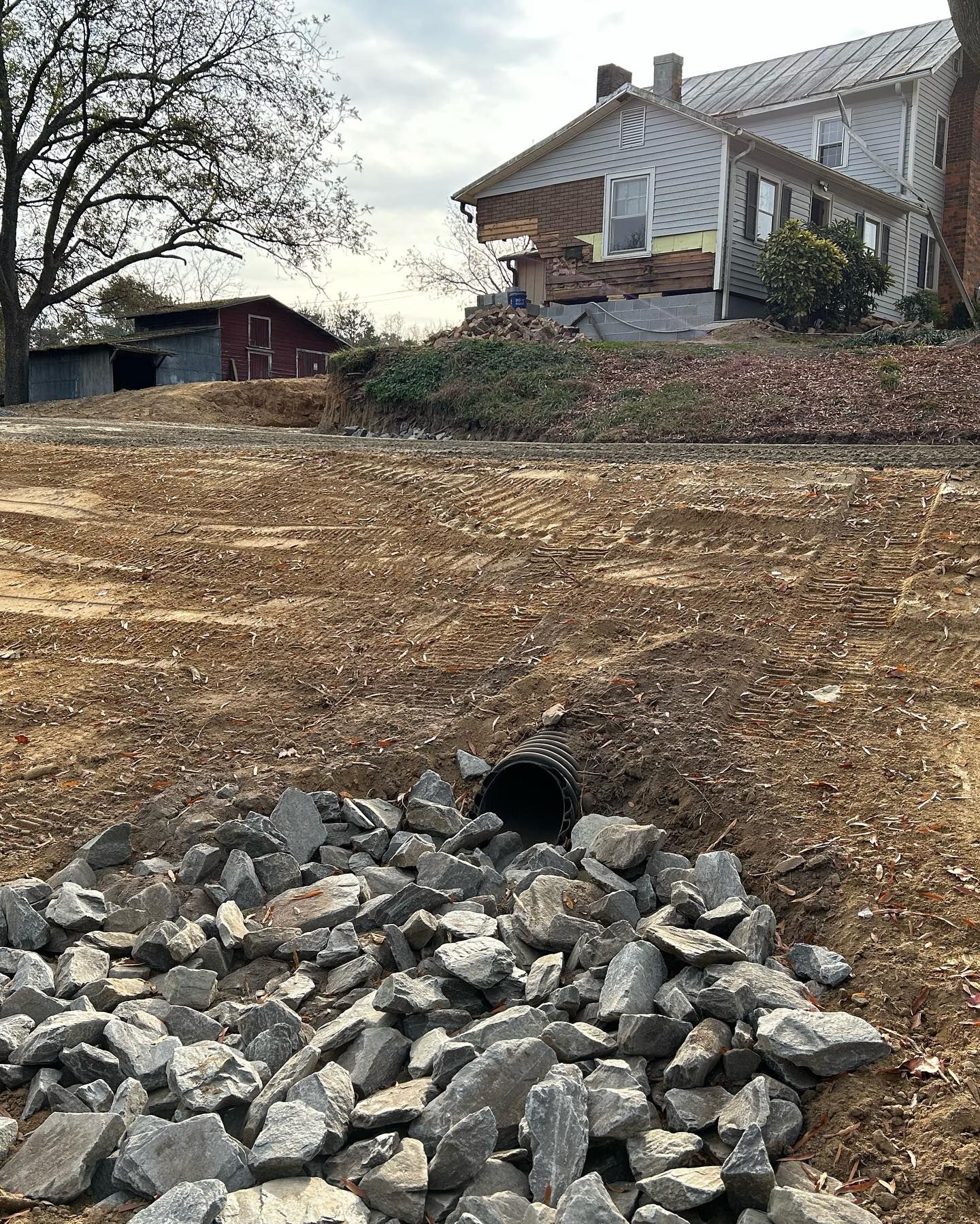 Rock-lined drainage pipe on a muddy slope below a white house and leafless trees