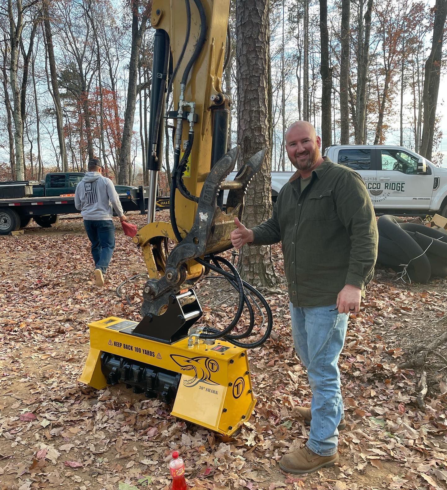 Man standing beside a yellow excavator attachment in a wooded worksite, with another person and truck in the background.