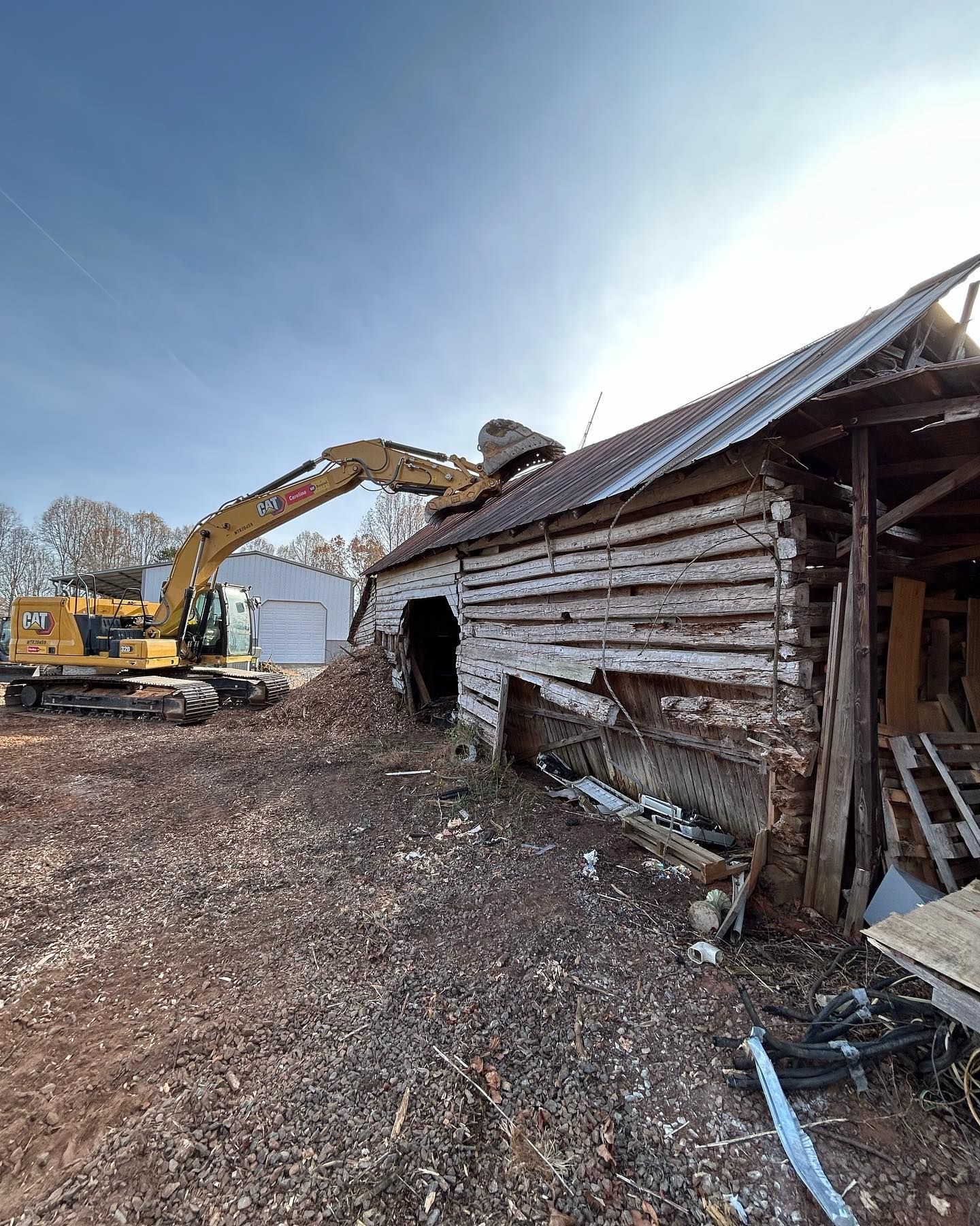 Excavator demolishing an old wooden barn amid debris under a clear blue sky