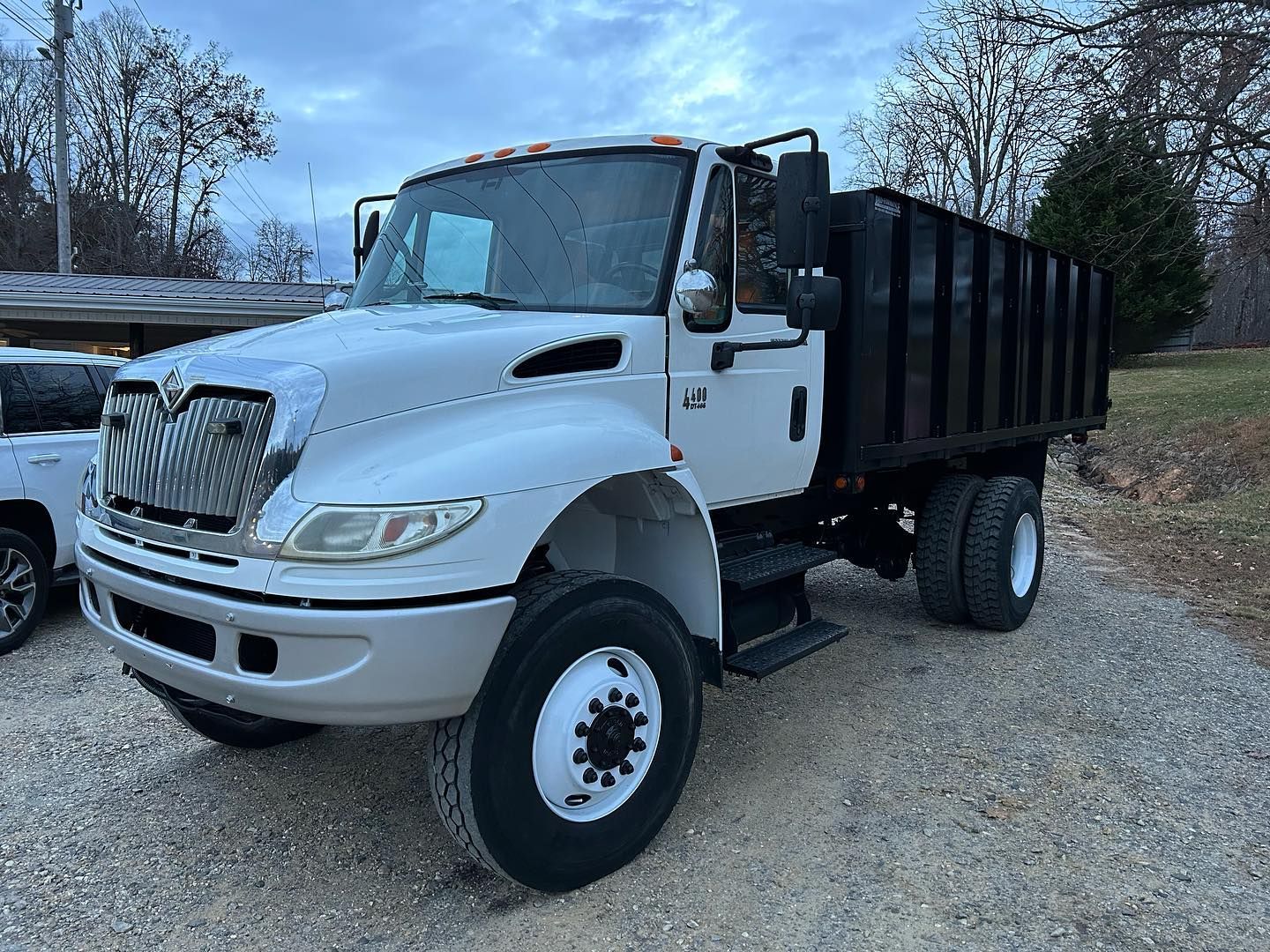 White dump truck parked on gravel, front-left view, with black cargo bed and trees in the background