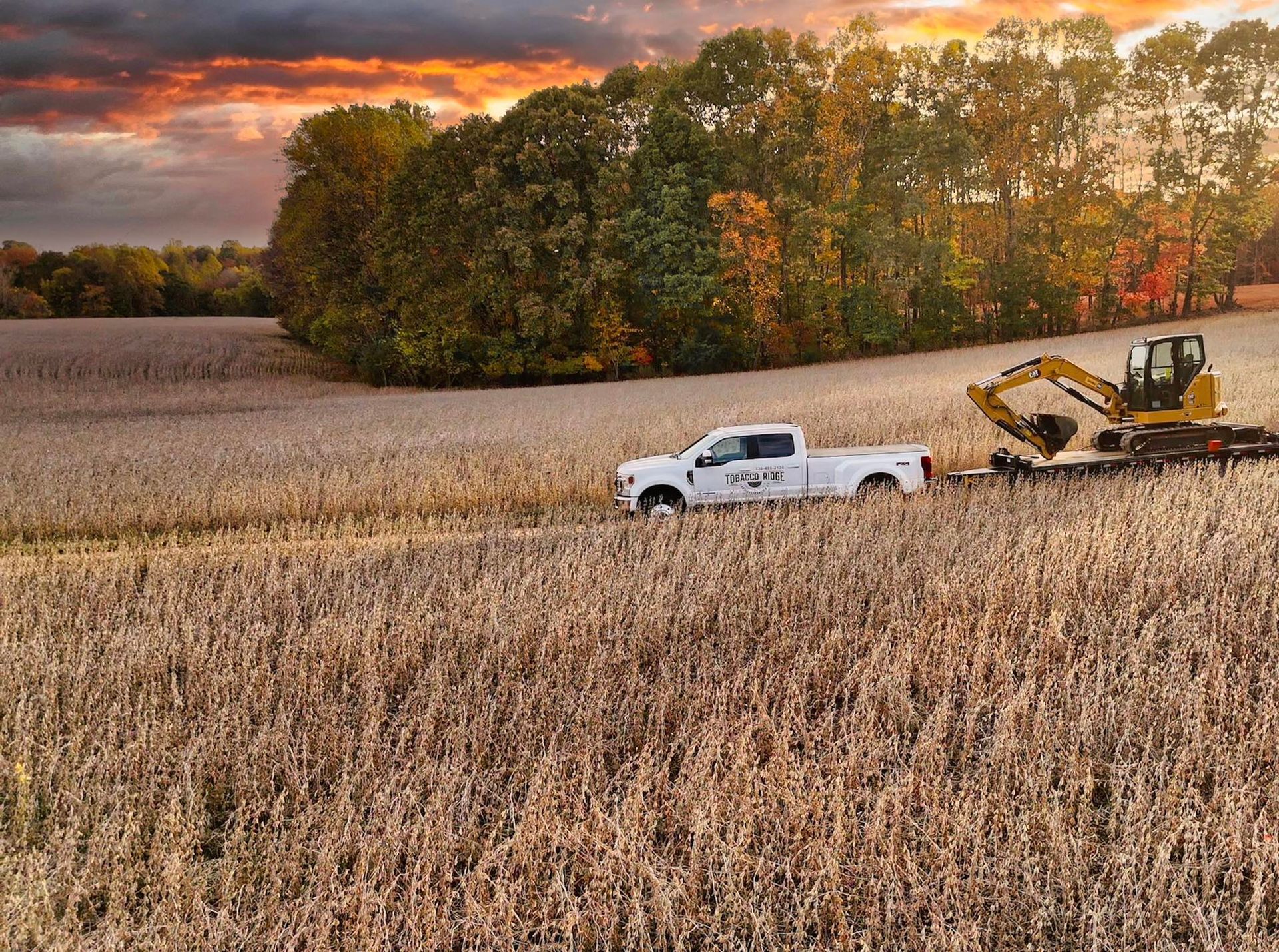 White pickup towing a yellow excavator through a harvested field at sunset