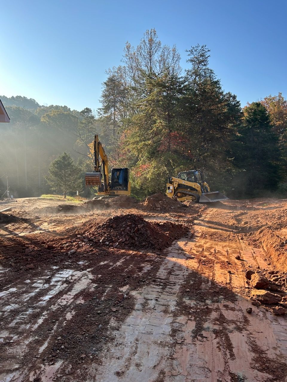 Construction site with excavator, dirt piles, and trees in warm morning light