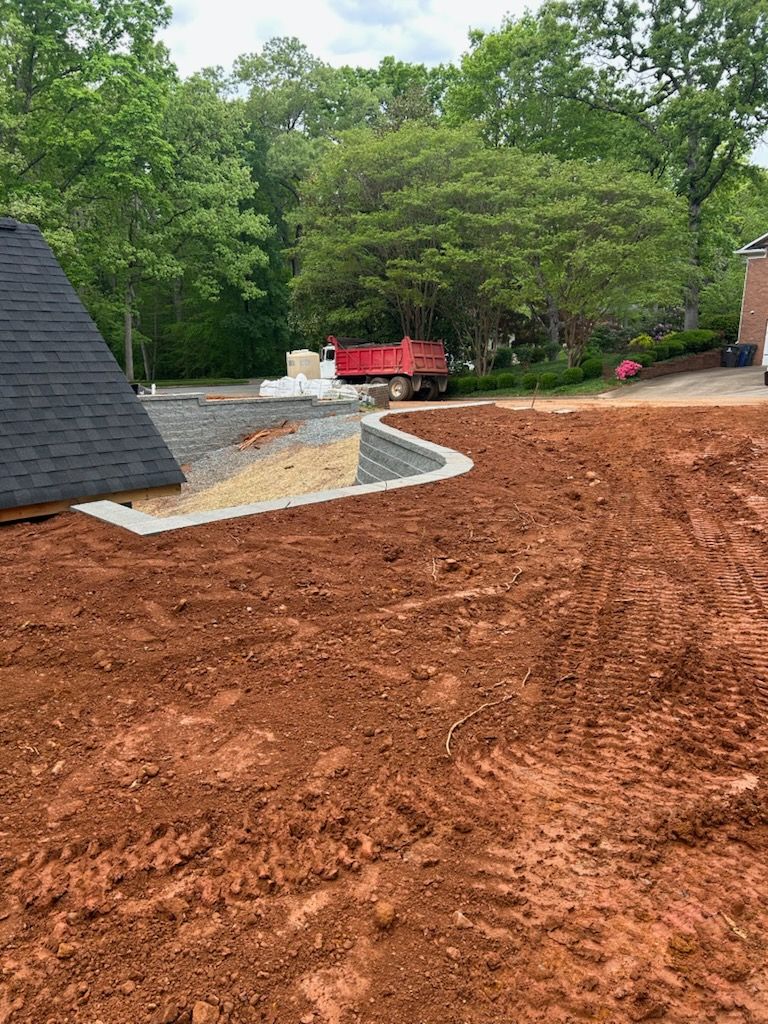 Freshly graded red dirt yard beside a house with a stone patio and trees in the background