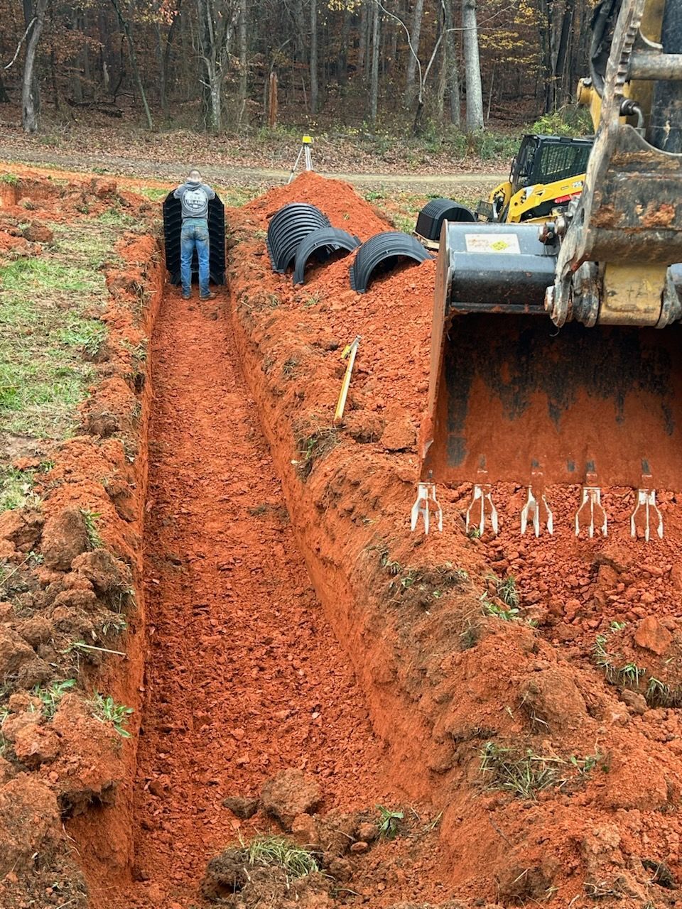 Excavator digging a trench with orange soil; black pipes and tools line the muddy excavation site.