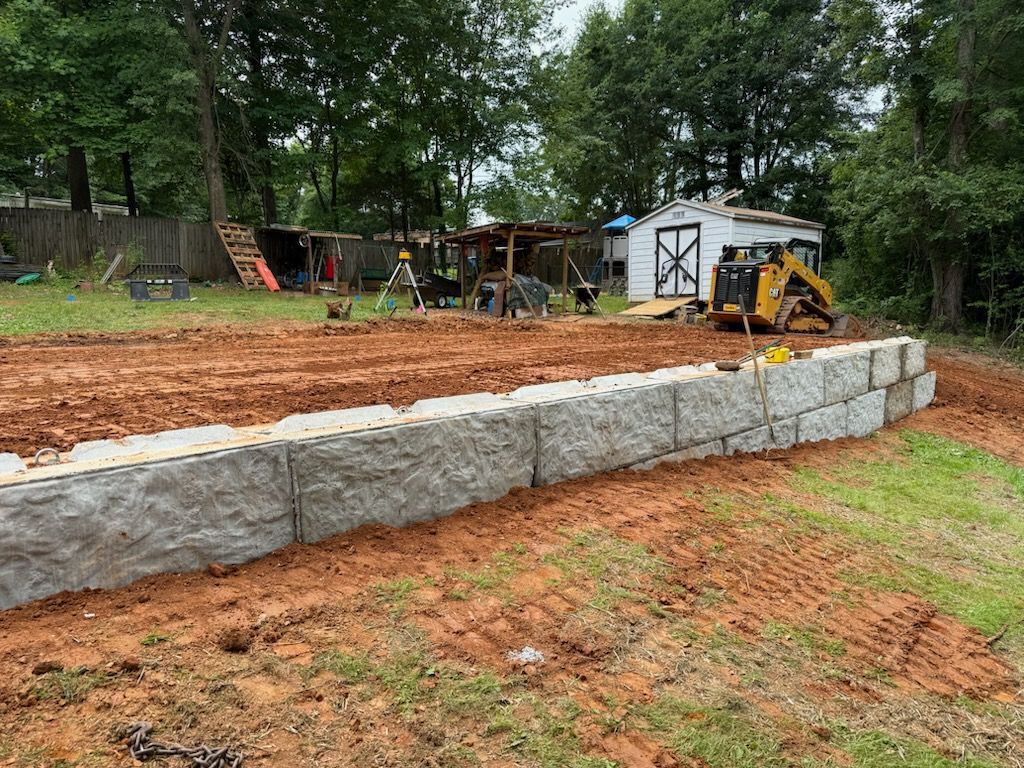 Stone retaining wall under construction beside a yard with a yellow skid steer and shed in the background