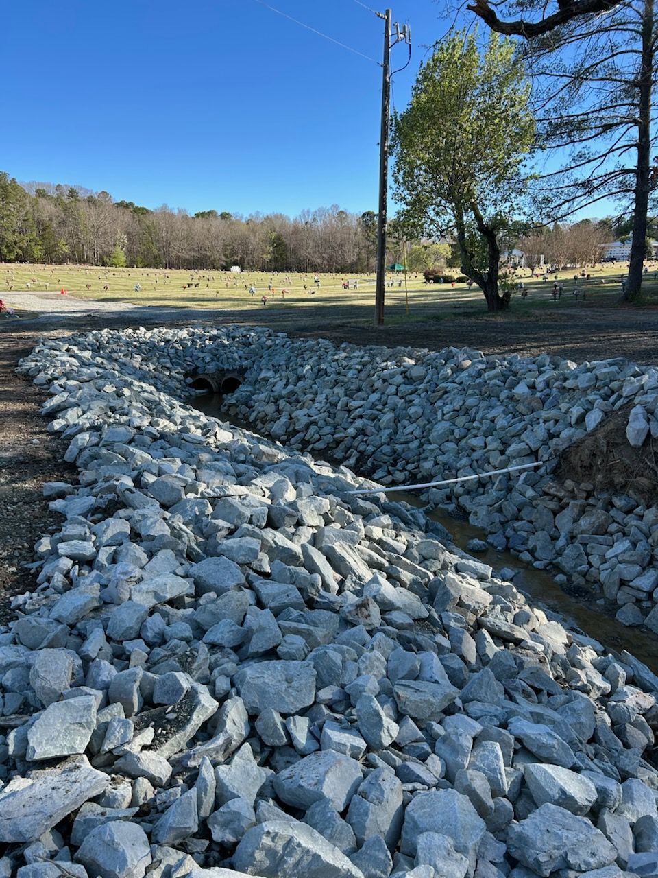 Rock-lined drainage ditch in a grassy field with trees and a utility pole under a clear blue sky