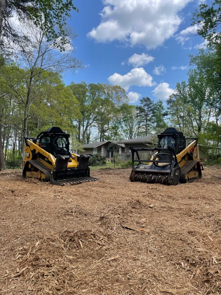 Two yellow skid-steer loaders in a wooded clearing under a blue sky with clouds
