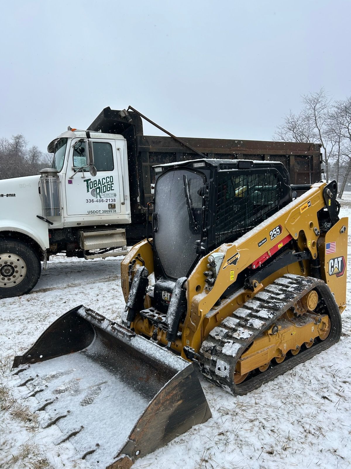 Yellow skid steer loader in snowy lot beside a white dump truck with raised bed