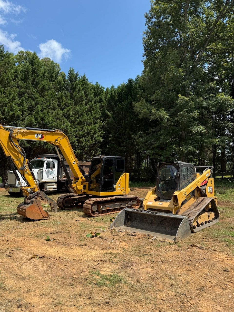 Yellow excavator and skid steer parked on a dirt lot beside trees under a blue sky.