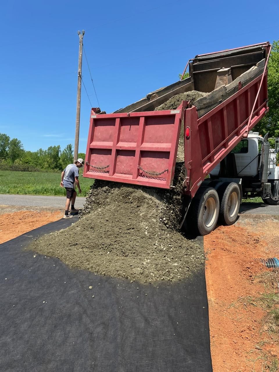 Dump truck unloading gravel beside a road, with a worker standing nearby under a clear blue sky
