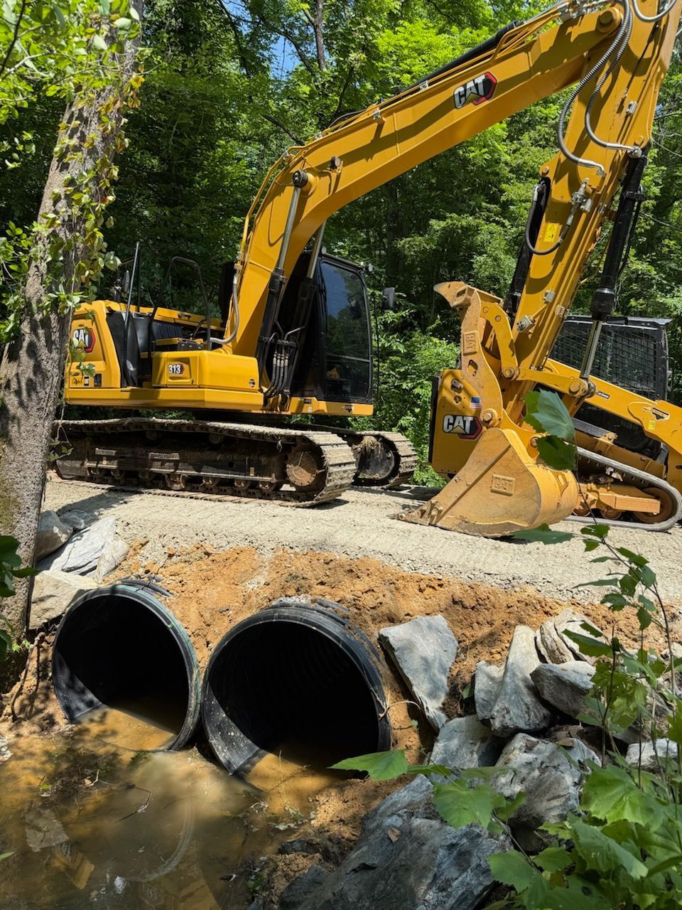 Yellow excavator working near twin culvert pipes in a wooded construction site