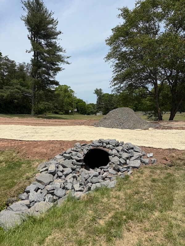 Small rock-lined pipe opening in a grassy park with a dirt path and trees in the background