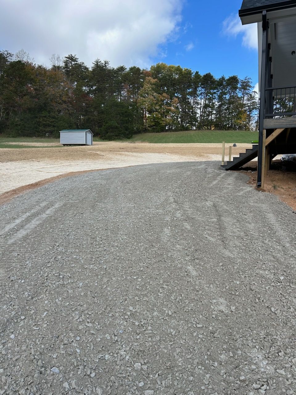 Gravel driveway beside a black building, with a grassy field and trees under a blue sky.