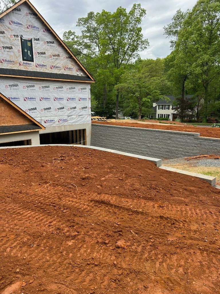 New house under construction with red dirt yard, gray retaining wall, and wooded background