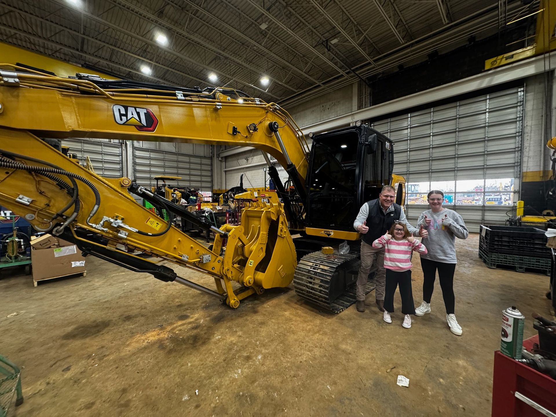 Two people posing beside a large yellow CAT excavator inside a warehouse-like garage