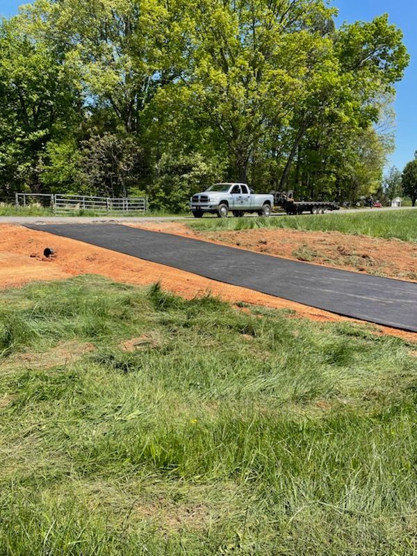 Rural roadside with a pickup truck beside a newly paved black path and grassy field under trees