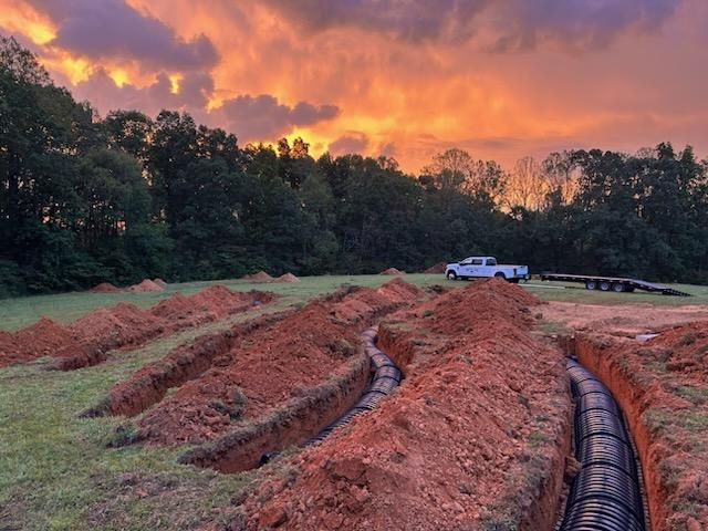Sunset over a trenching site with red dirt rows, pipes, trees, and a white truck in the distance