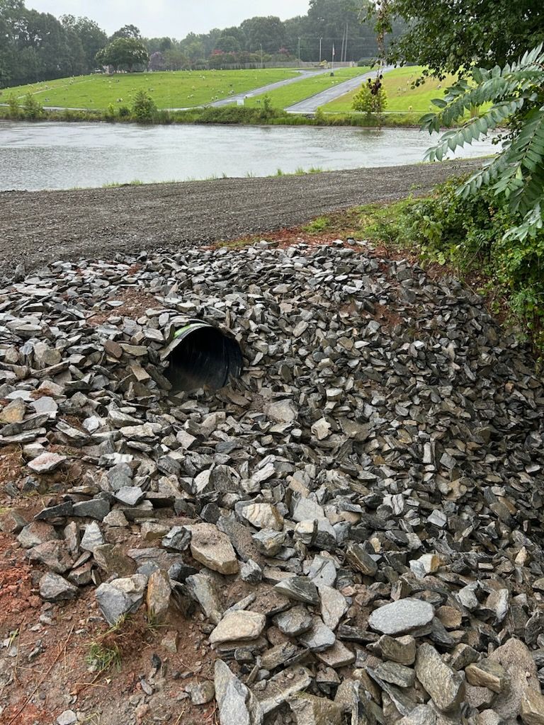 Rock-lined drainage channel with a culvert pipe beside a pond and grassy park lawn