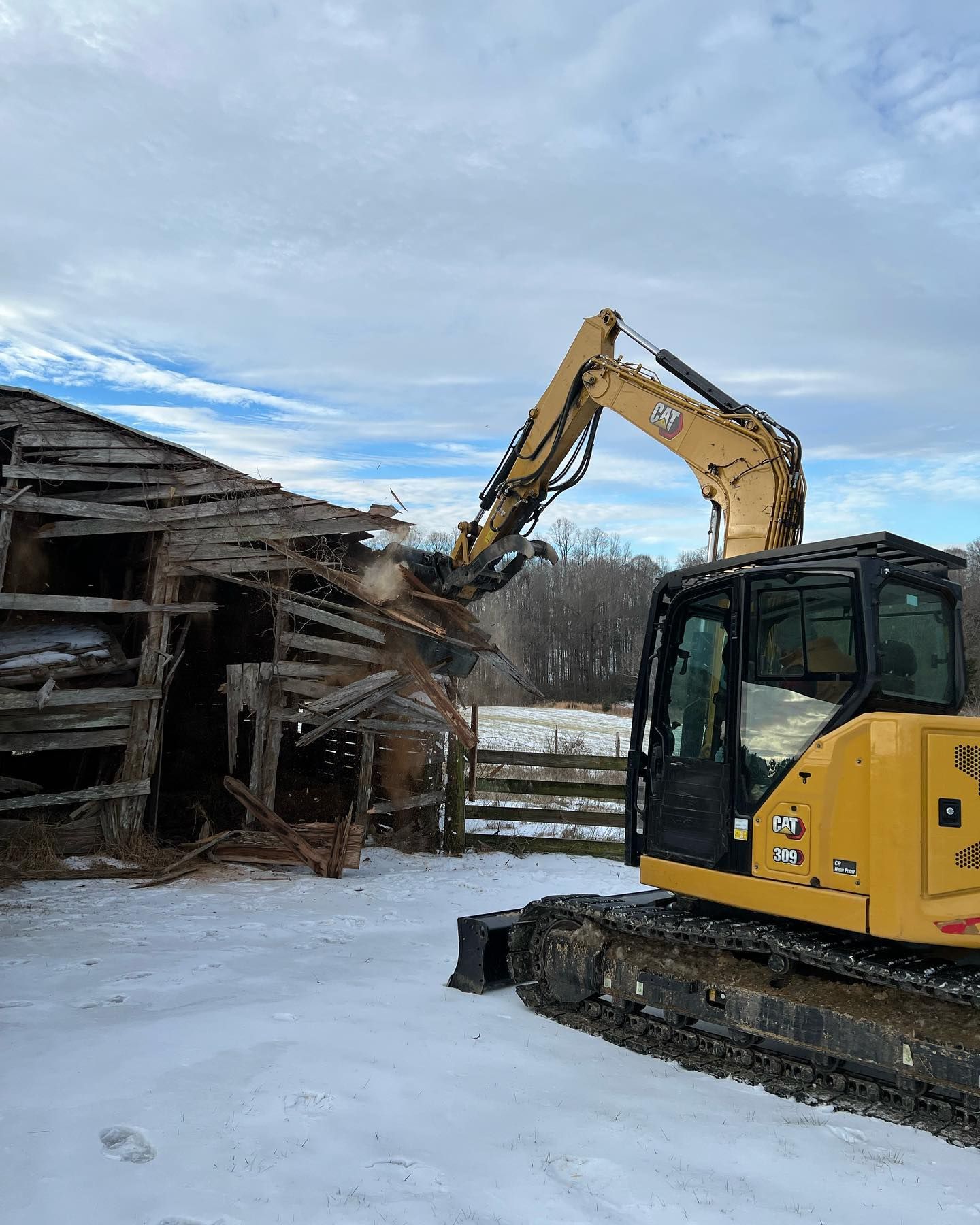 Yellow excavator demolishing a weathered wooden barn in a snowy field