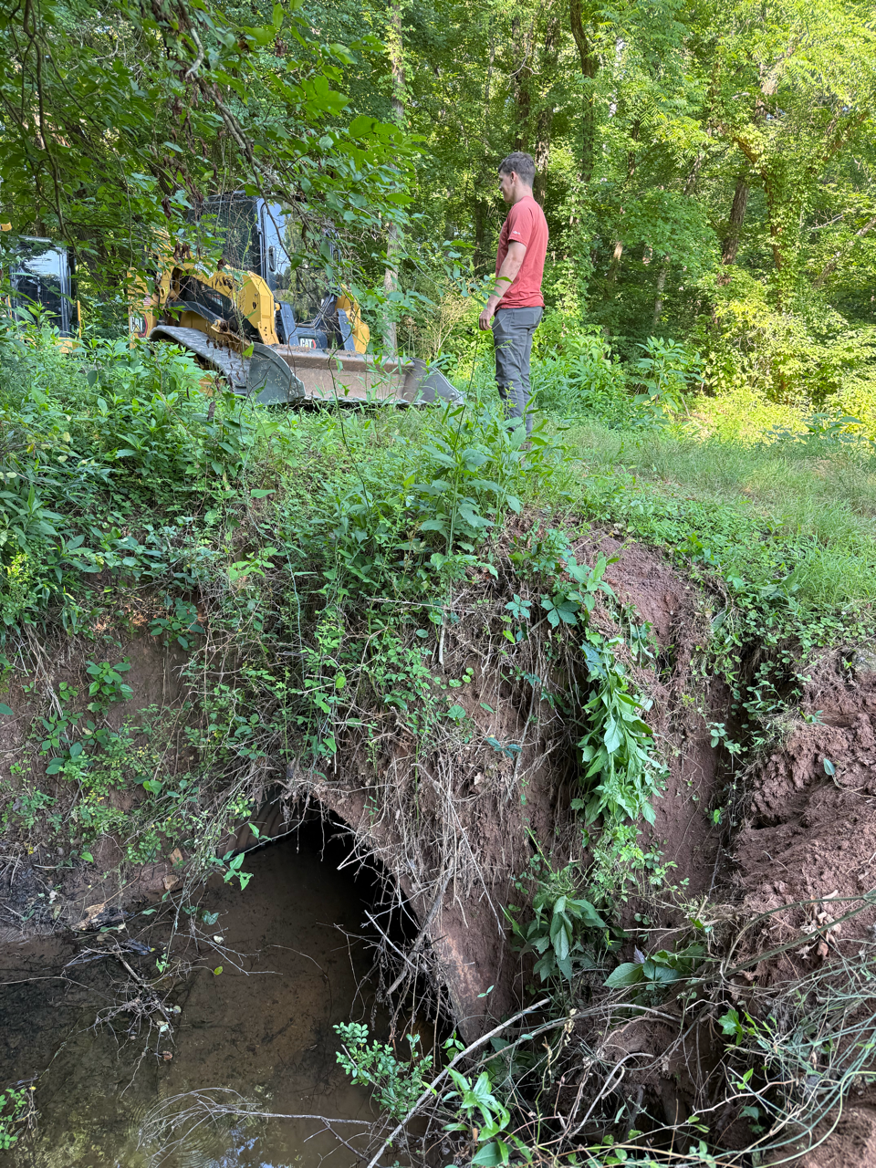 Person in red shirt stands by a backhoe on an eroded stream bank in a wooded area.