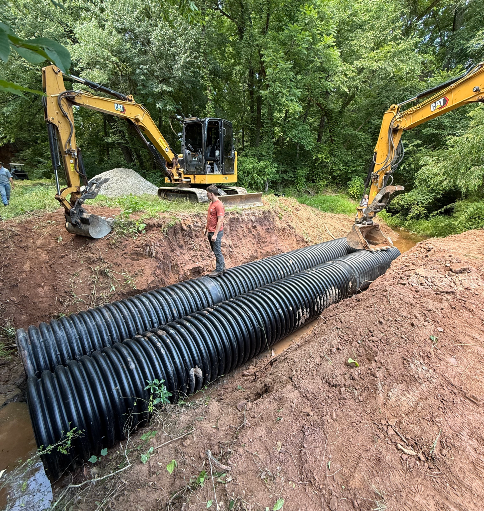 Two excavators install a large black corrugated pipe in a muddy trench surrounded by trees.