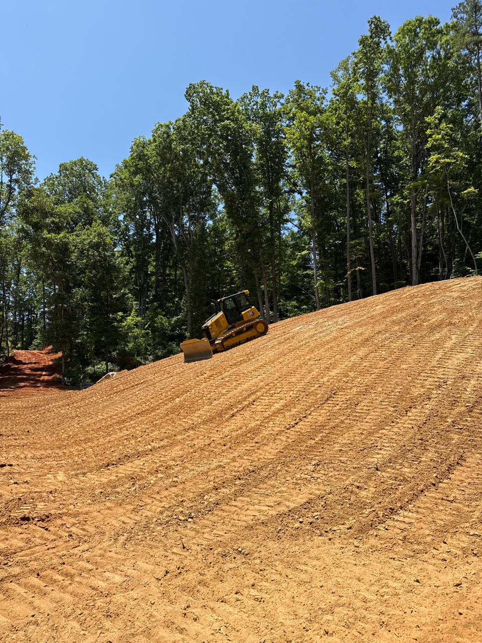 Yellow bulldozer grading a steep dirt slope beside a forest under a clear blue sky