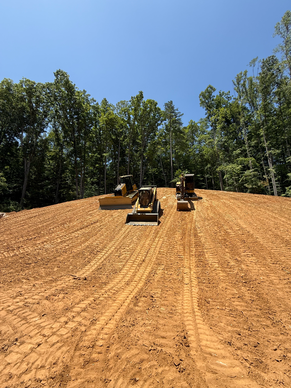 Three bulldozers grading a dirt slope beneath a clear blue sky, with trees lining the background.