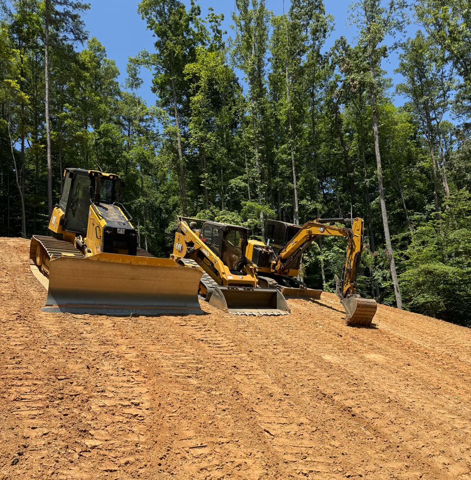 Two yellow excavators and a bulldozer on a cleared dirt slope in a forest