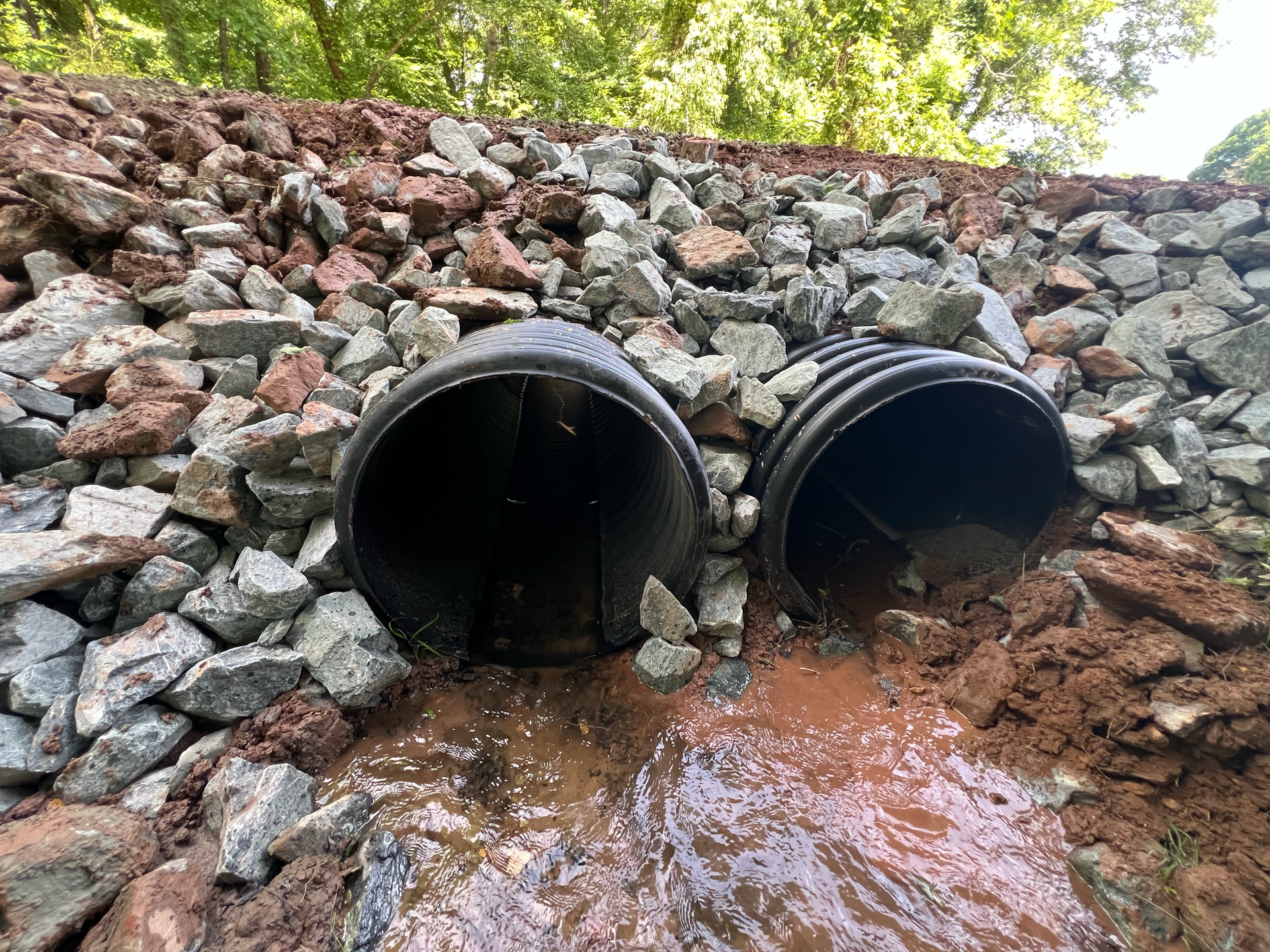 Two black drainage pipes in a rocky ditch, with water flowing out beneath a leafy embankment