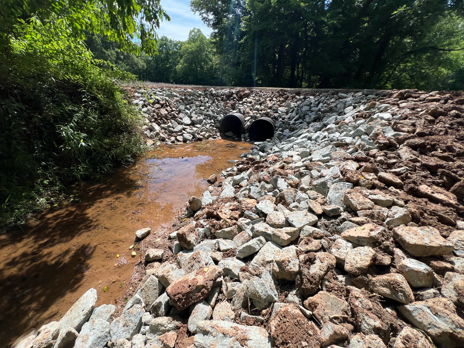 Rock-lined drainage ditch with twin culverts under a stone embankment, surrounded by trees.