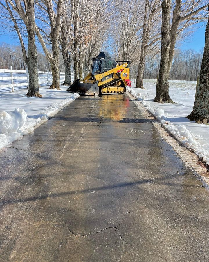 Yellow tractor clearing snow from a paved path lined with bare trees on a winter day