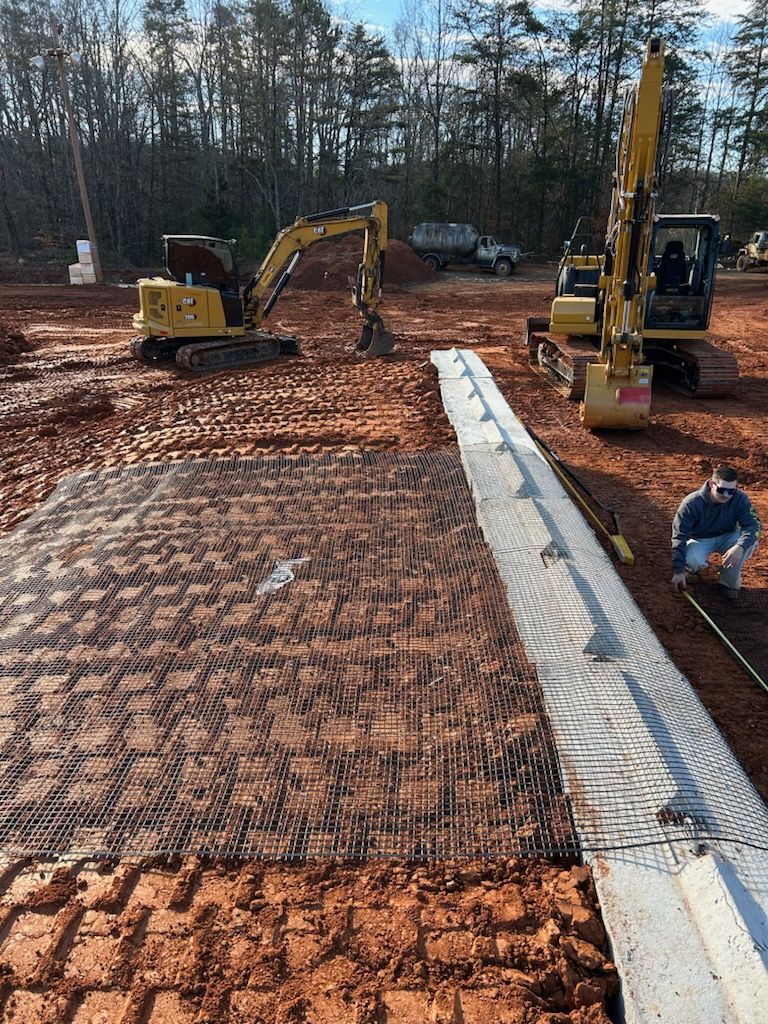 Construction site with two excavators grading red dirt beside a concrete curb and metal reinforcement mat.
