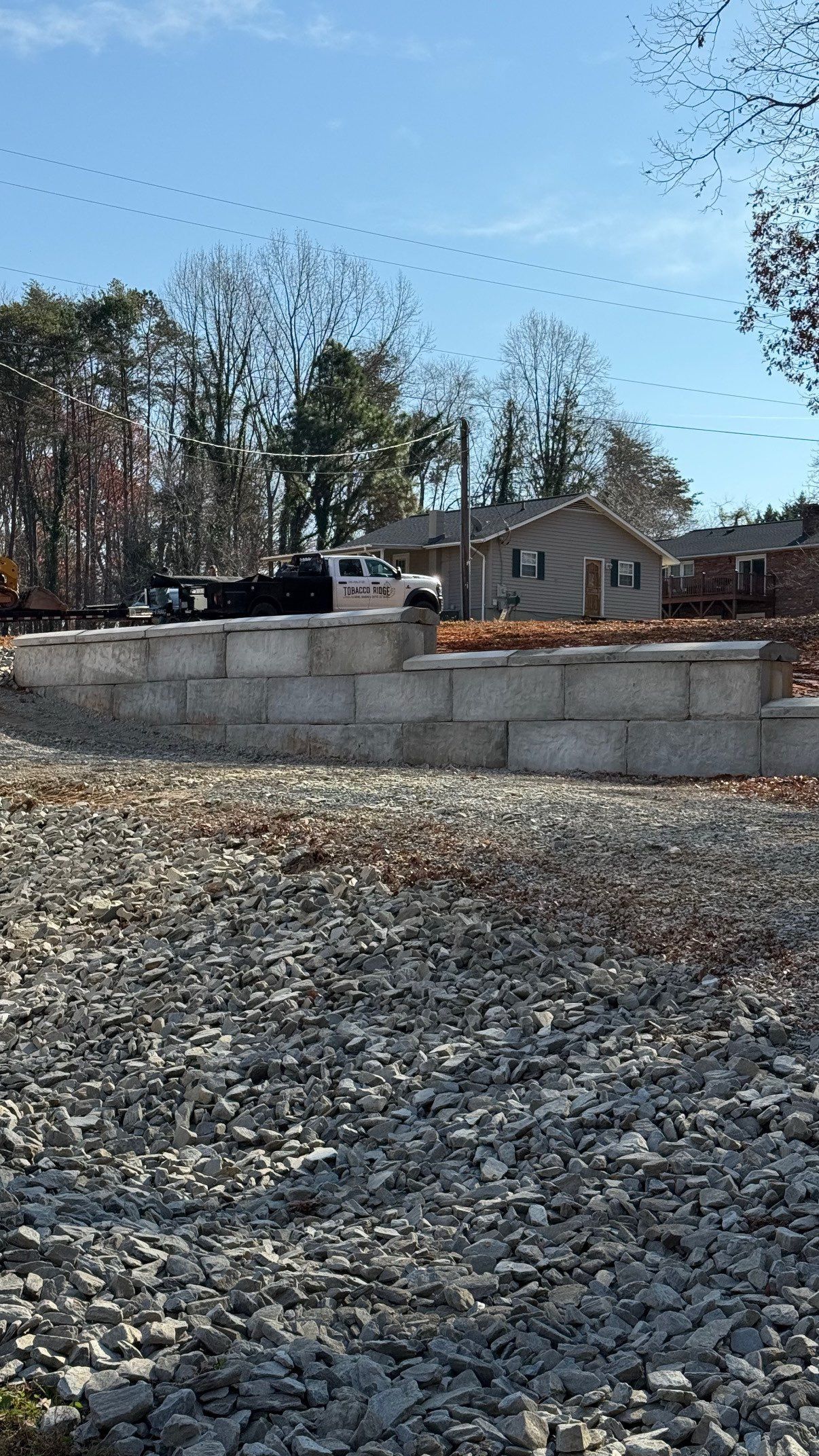 Gravel lot with a low stone wall, bare trees, and a house under a clear blue sky